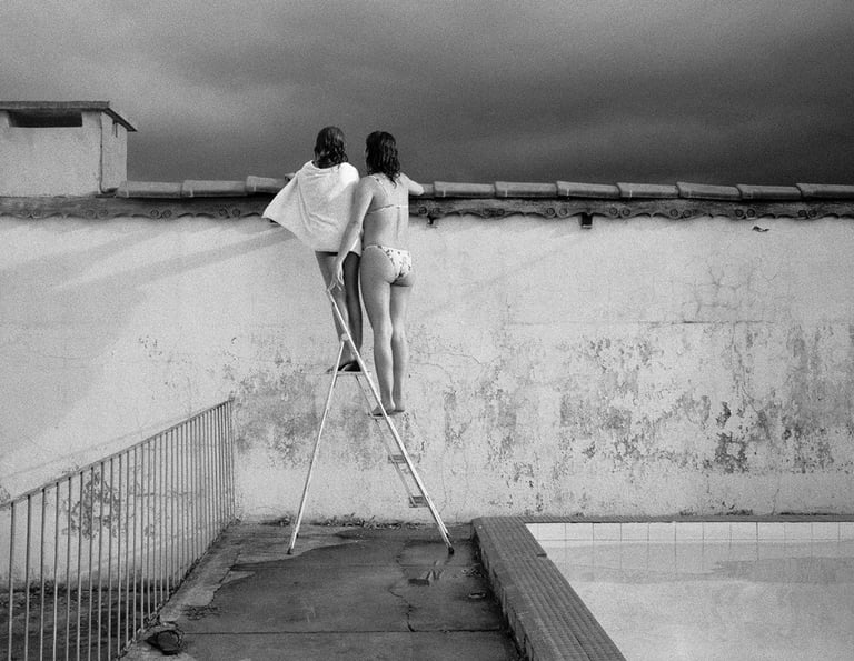 Duas mulheres de costas em cima de escada apoiada em muro, observando o horizonte sob céu nublado.