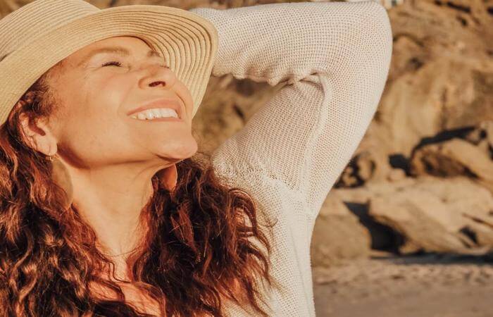 Person with long hair and woven hat smiling with eyes closed, standing near a sunlit rocky shore.