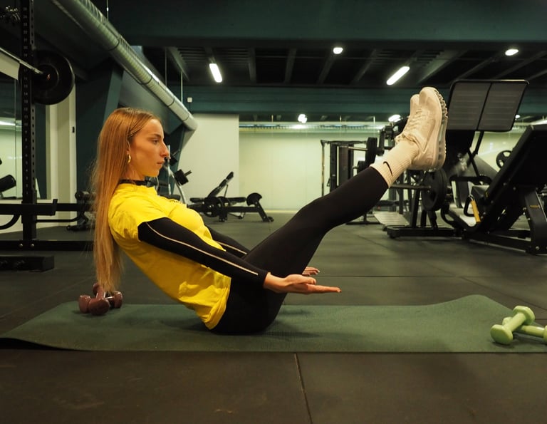 Una donna in maglia gialla esegue l'esercizio 'boat pose' su un tappetino da yoga in palestra, concentrandosi sull'equilibrio