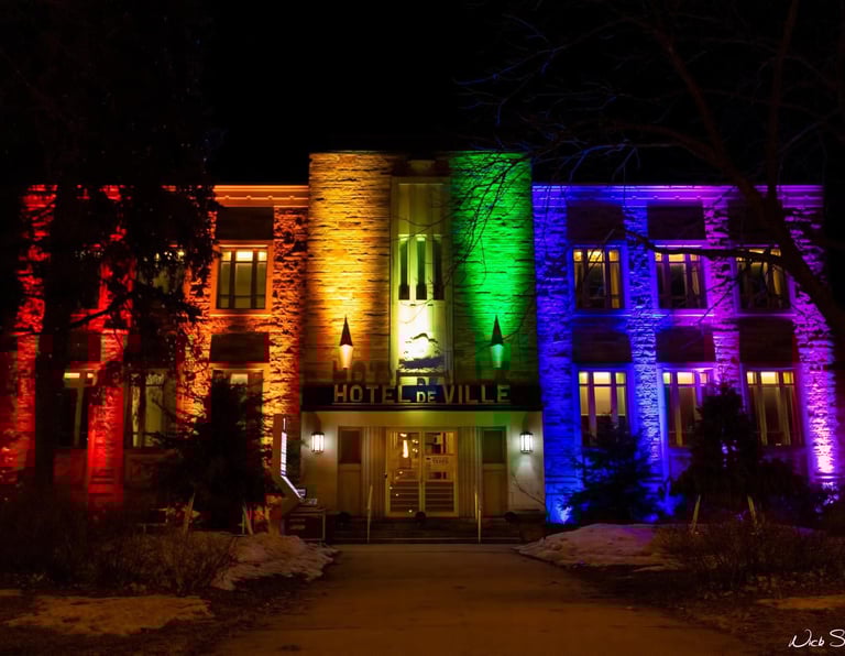 The Hotel de Ville building illuminated with vibrant rainbow pride colors at night.