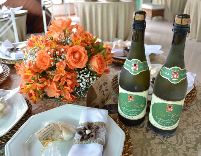 "Mesa de casamento decorada com arranjo floral de rosas laranjas e gipsofila, garrafas de Lambrusco 