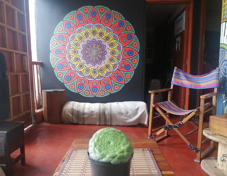 Sala con planta y mandala pintado en la pared, hay una silla mecedora y otros muebles rústicos con luz cálida