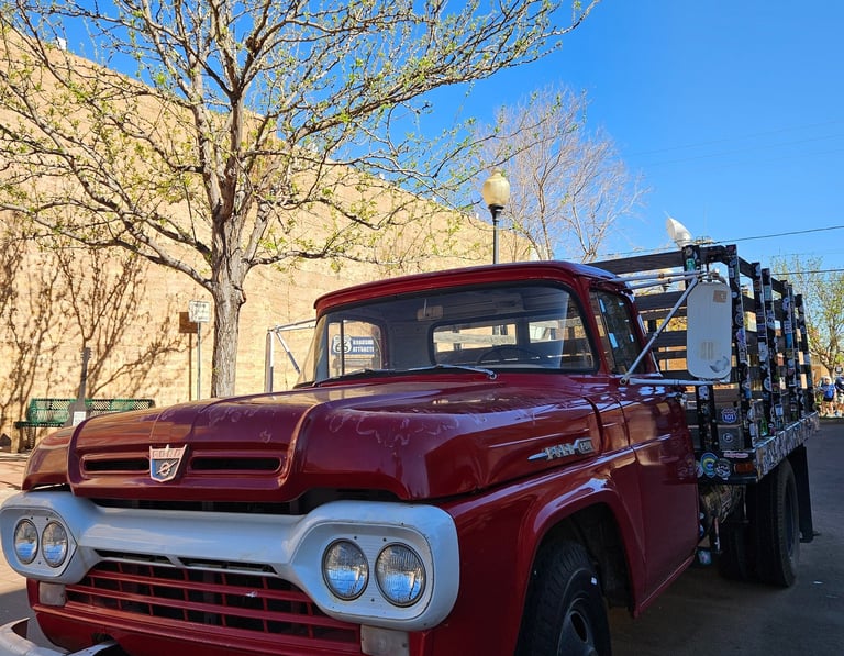 Classic red Ford flatbed truck parked on a sunny city street with budding spring trees.