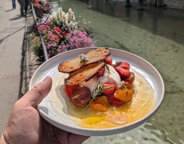 Une personne tient une assiette avec une burratta accompagné de fraises, tomates et un croustillant