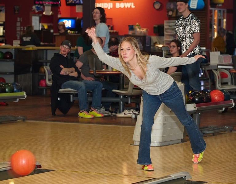 A blonde woman in jeans throws an orange bowling ball down the lane at a local bowling alley.
