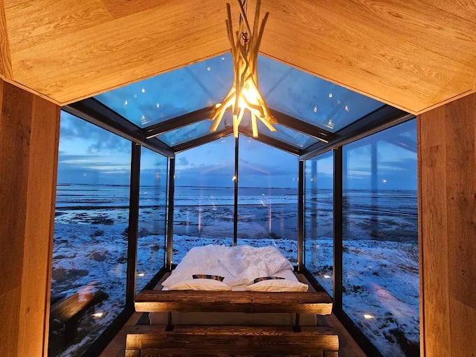 A modern glass cabin bedroom in Iceland with panoramic views of a snowy coastal landscape at dusk.