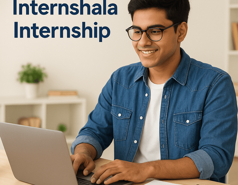 A smiling young South Asian student wearing glasses and a denim shirt works on a laptop at a desk wi