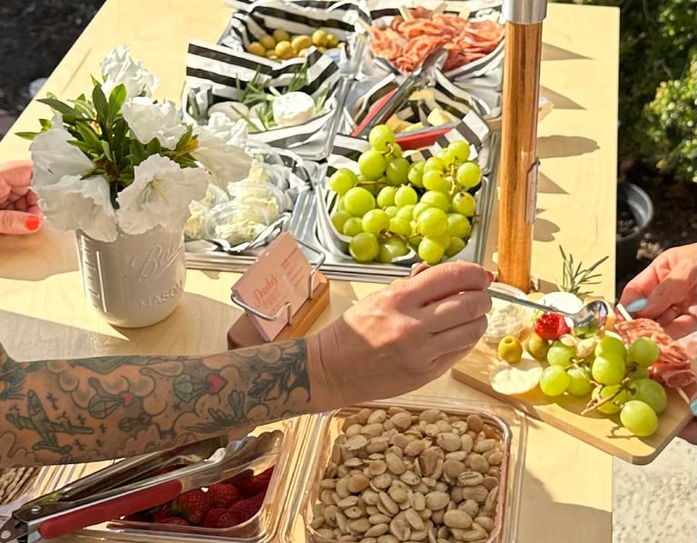 a person putting topping on a charcuterie board on a bar cart with charcuterie times in the backgrou