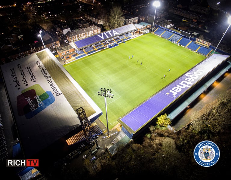 Aerial night view of Stockport County FC Edgeley Park stadium illuminated by bright floodlights.