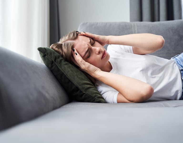 A woman lying on a grey sofa holding her head due to a severe migraine or tension headache.