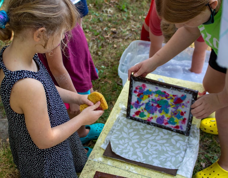 Group of young children holding a colorful screen above a piece of fabric while making paper.