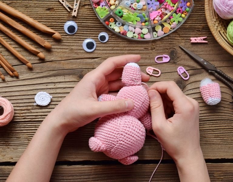Manos ensamblando un amigurumi rosa tejido a crochet, rodeado de ganchos y estambre.