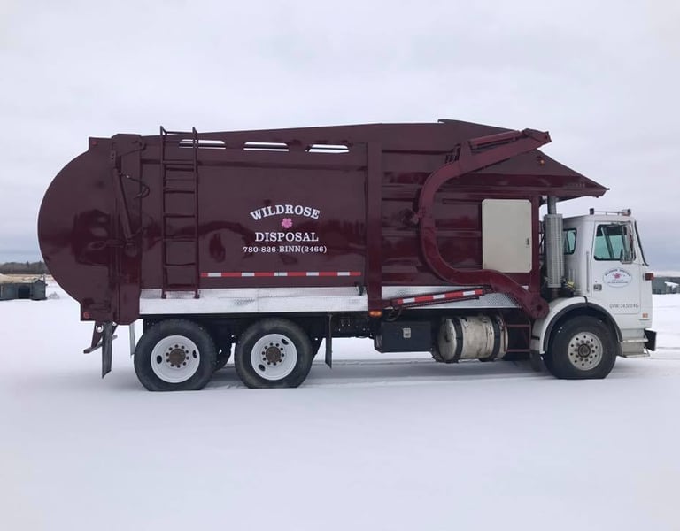 A maroon Wildrose Disposal front-load garbage truck parked in a snowy field for waste management services.