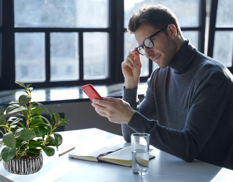 A man in glasses looks at his red smartphone at a bright desk with a potted green plant.