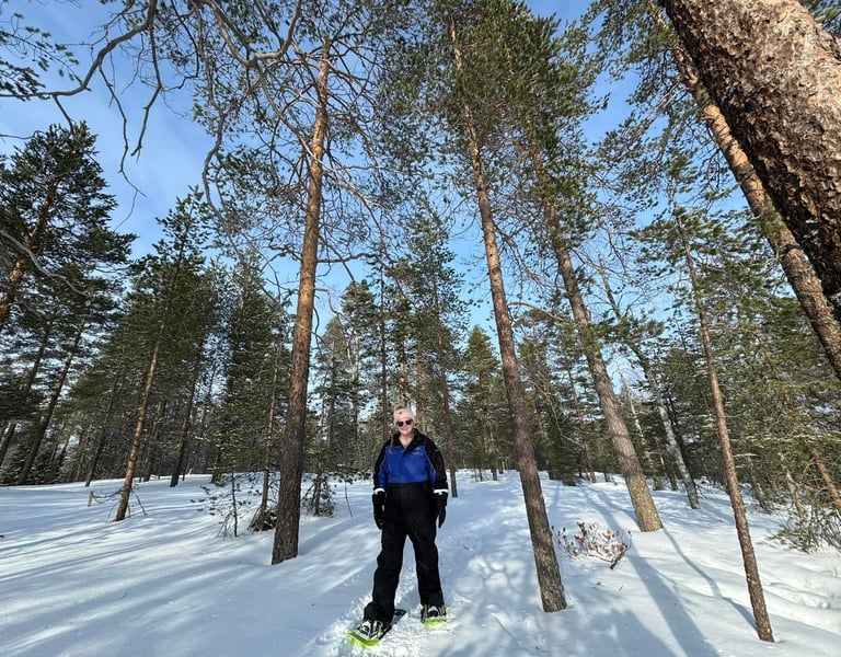 A woman in winter gear snowshoeing through a sunny, snow-covered pine forest in Lapland.