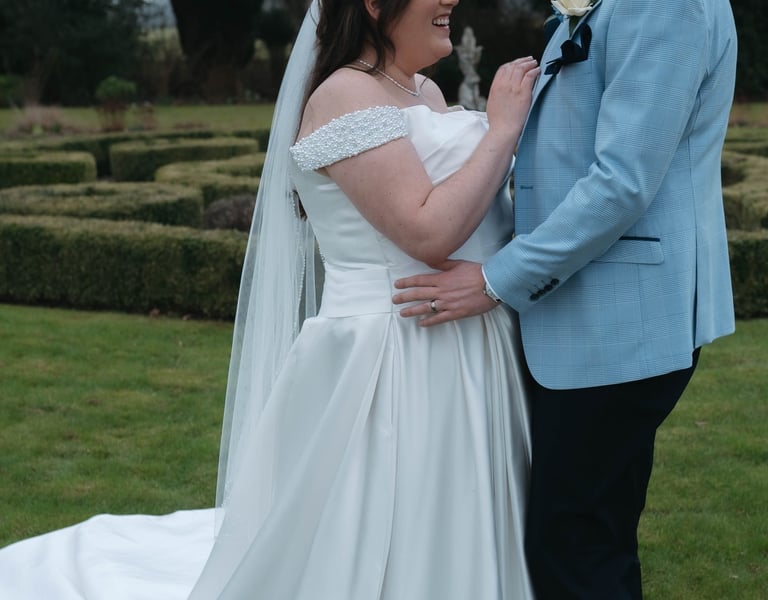 A smiling bride in a white off-the-shoulder wedding dress poses with her groom in a blue suit jacket.