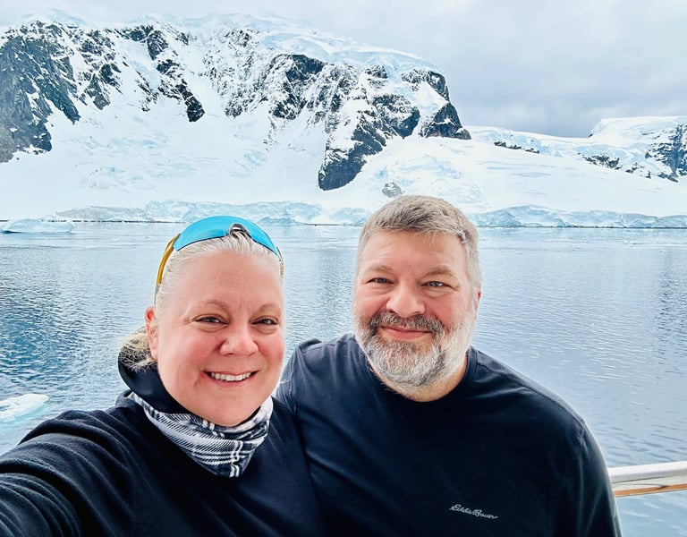 Smiling couple taking a selfie on an Antarctica cruise with snowy mountains and glaciers in the background.