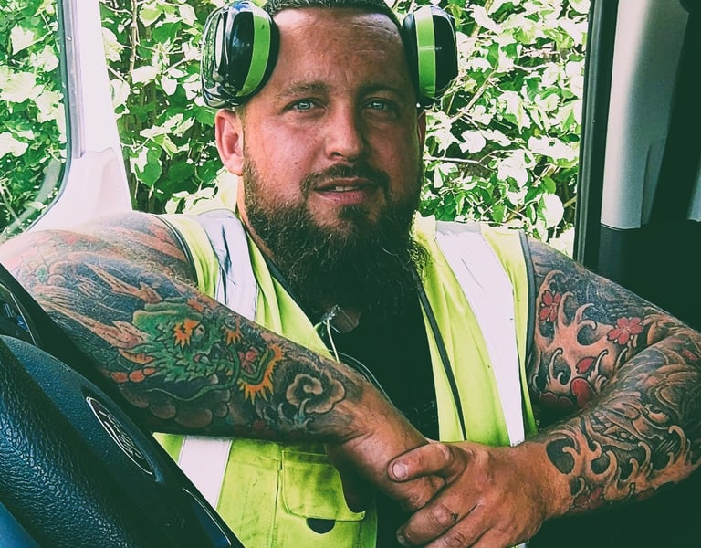 Tattooed construction worker wearing a high-visibility vest and ear protection sitting in a van.