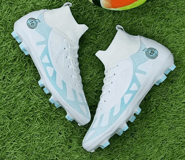 Botines de fútbol de caña alta en blanco y azul claro con tacos para terreno firme sobre césped.