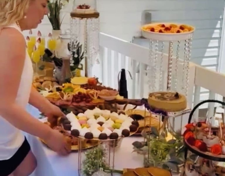 Una mujer organiza un buffet de brunch gourmet con postres, pasteles y aperitivos en un balcón.
