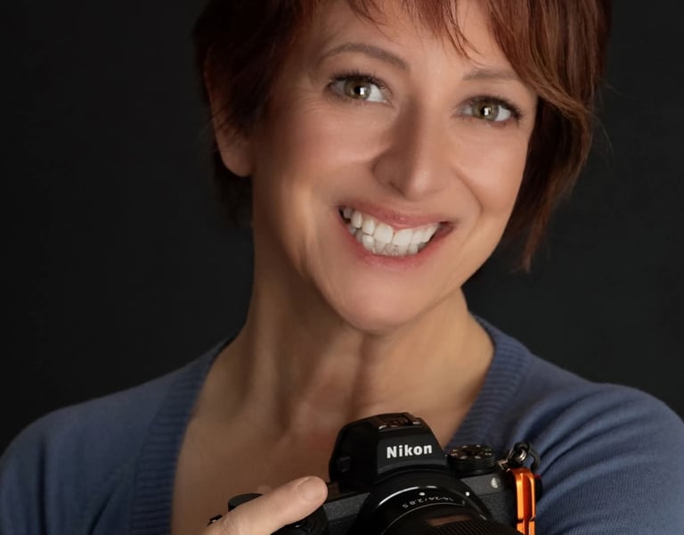 A smiling woman with short auburn hair holding a professional Nikon mirrorless camera.