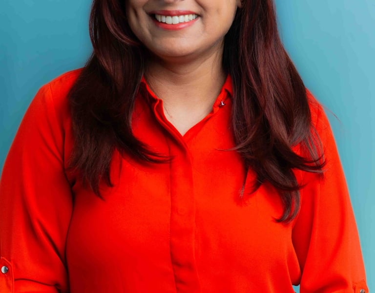 A smiling woman in glasses stands against a blue background, wearing a vibrant red shirt and a friendly demeanor.