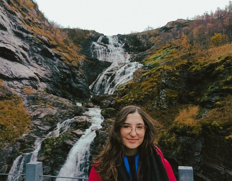A smiling young woman in a red jacket posing in front of a majestic waterfall in Norway.
