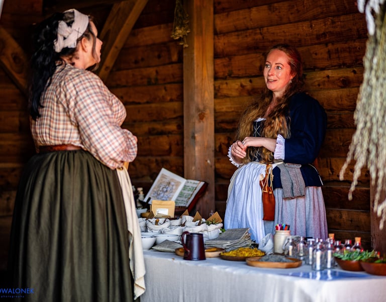 a woman in 18th century clothing stands behind a table covered in herbal remedies