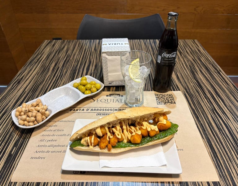 Spanish bocadillo sandwich with fried calamari and green sauce served with olives, peanuts, and a glass of ice.