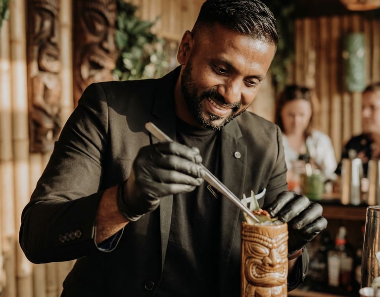 Smiling professional bartender garnishing a tropical cocktail in a ceramic tiki mug at a bamboo bar.