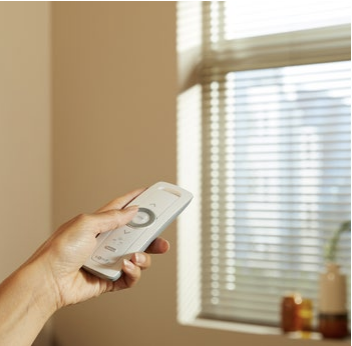 Hand using a remote control to operate motorized window blinds in a bright room.