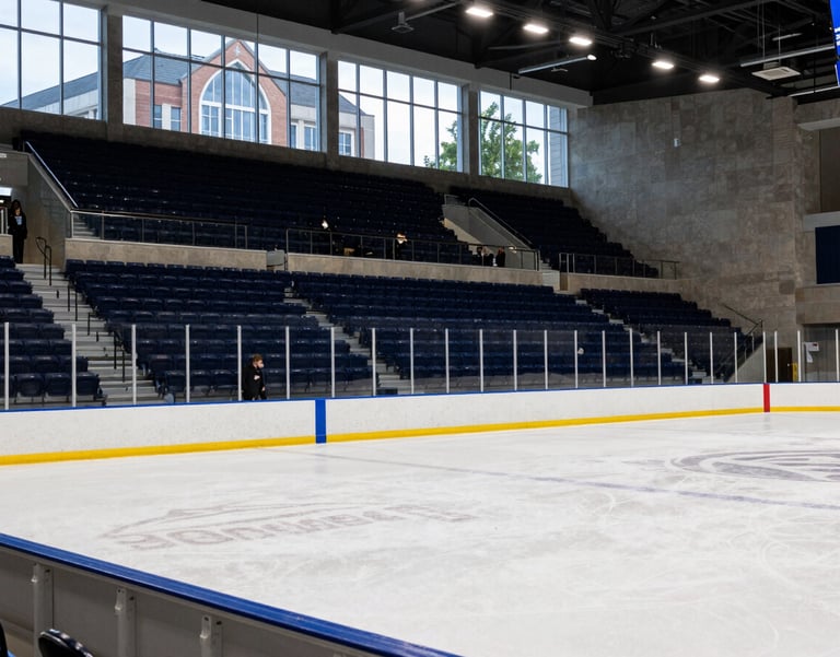A crisp, wide-angle shot of a modern collegiate ice hockey rink in a North American / US Southern university campus setting. The lighting is bright and professional, highlighting the dark navy blue and royal blue accents of the stadium seating. In the background, students walk past large glass windows showing the architectural style of a Southern educational institution.
