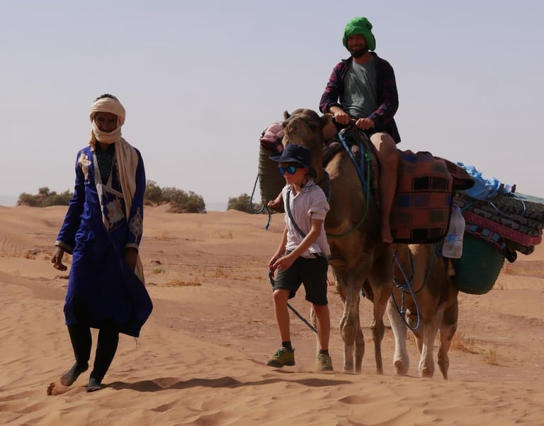 A boy leads a man riding a camel on a Sahara desert trek, while a nomad guide walks in front