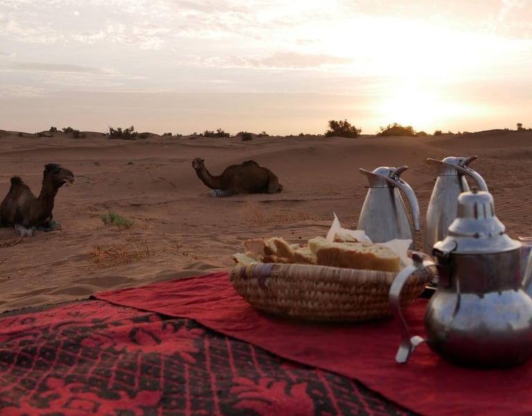 Teapots and a basket of bread laid out for tea in the desert, as two camels look on