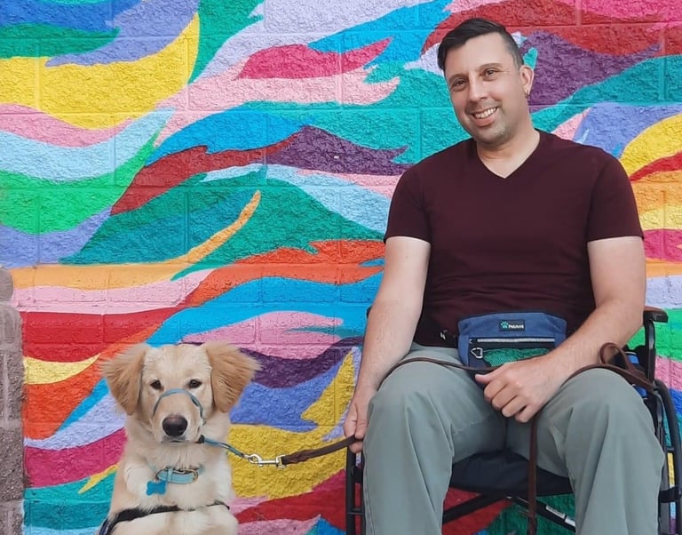 A man in a wheelchair with his golden retriever service dog in front of a colorful mural.