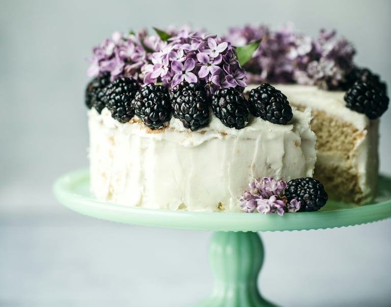 Pastel de vainilla en capas con betún blanco, decorado con moras frescas y flores lilas moradas.