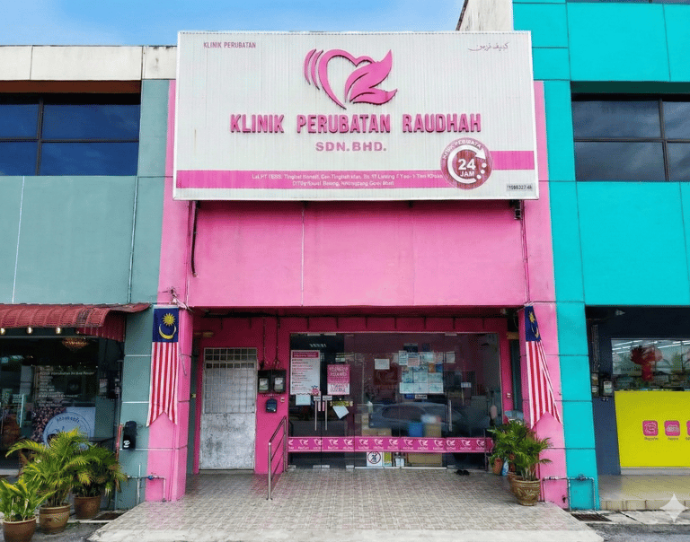 Pink storefront of Klinik Perubatan Raudah, a 24-hour medical clinic in Malaysia with glass doors and flags.