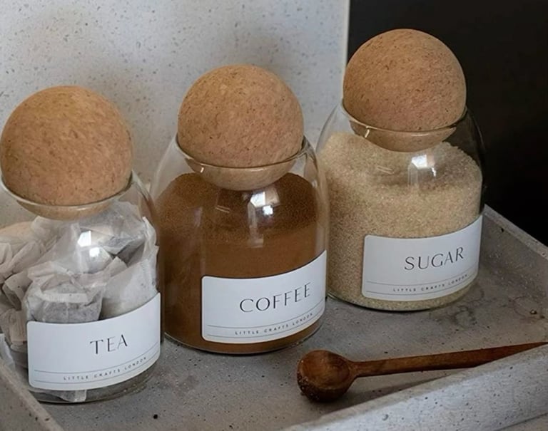 Minimalist glass storage jars for tea, coffee, and sugar with cork ball lids on a concrete tray.