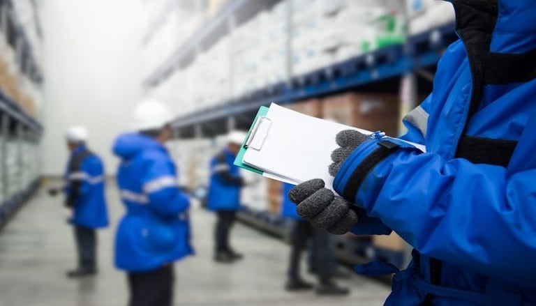 Warehouse worker in a blue winter jacket holding a clipboard for inventory management.