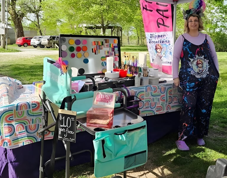 A table set up at a festival, colorful paints displayed with Sarah kipper in front of table smiling 