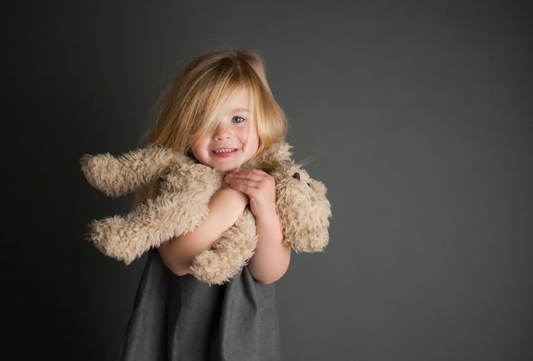 une petite fille blonde souriante faisant un calin / tenant un ours en peluche sur fond noir