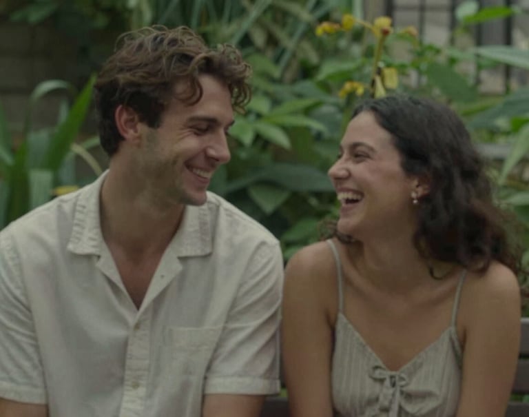 A candid moment of a couple sharing a quiet laugh in a lush green garden. The style is cinematic and story-driven, with subtle earth tones like #8F6E5F in the shadows.