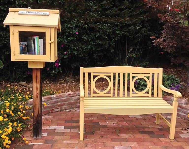 Yellow Little Free Library box on a post next to a matching yellow wooden garden bench on a brick patio.
