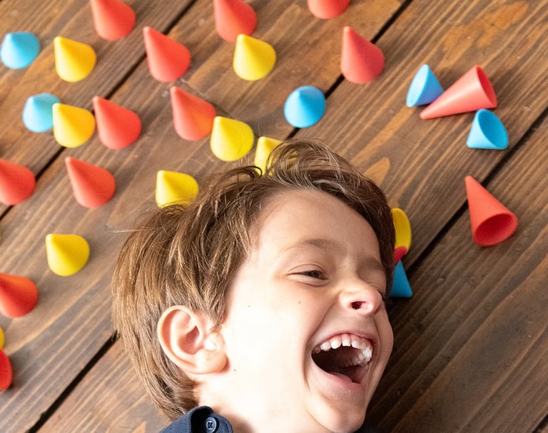 Laughing boy laying on the brown ground surrounded by blue, red and yellow silicone cones