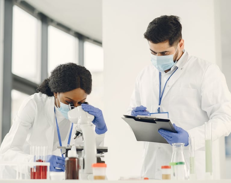 Woman in lab coat sitting looks into  a microscope while a man in lab coat stands with chart holder