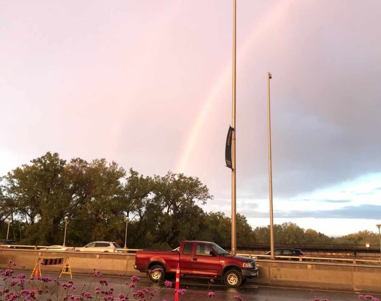 Red truck on a bridge with a beautiful rainbow in the sky, there are trees and flowers