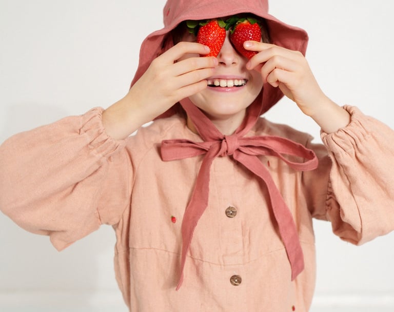 Smiling child in a dusty pink bonnet and dress playfully holding two strawberries over their eyes, d
