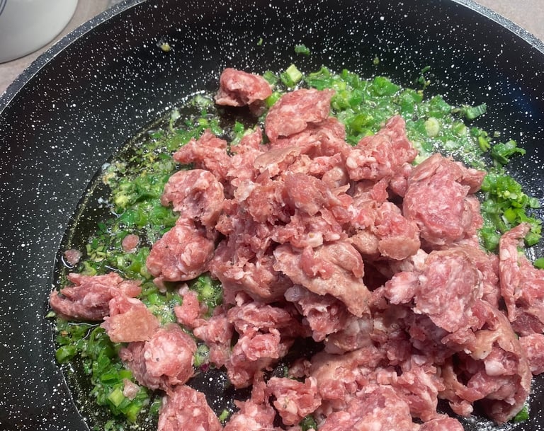 Raw ground sausage browning in a skillet with sautéed green onions and oil.