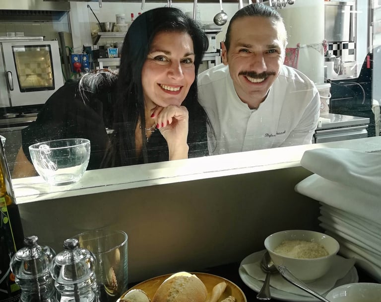 Clara Mennella e uno chef affacciati al pass della cucina, tra pane fresco e mise en place, simbolo del racconto culinario.