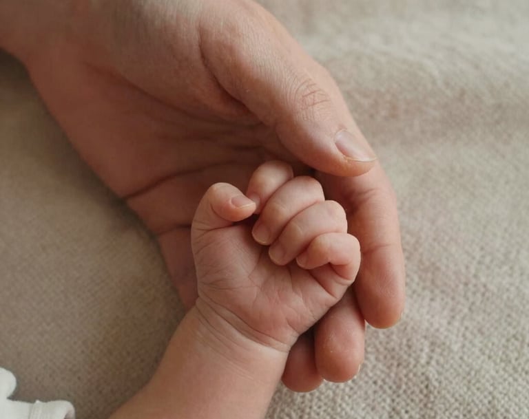 A detailed close-up shot of a parent's hand gently holding a newborn's tiny hand, set against a soft, light beige knit blanket. Captured in North American / US (West Michigan) with warm, natural morning light.